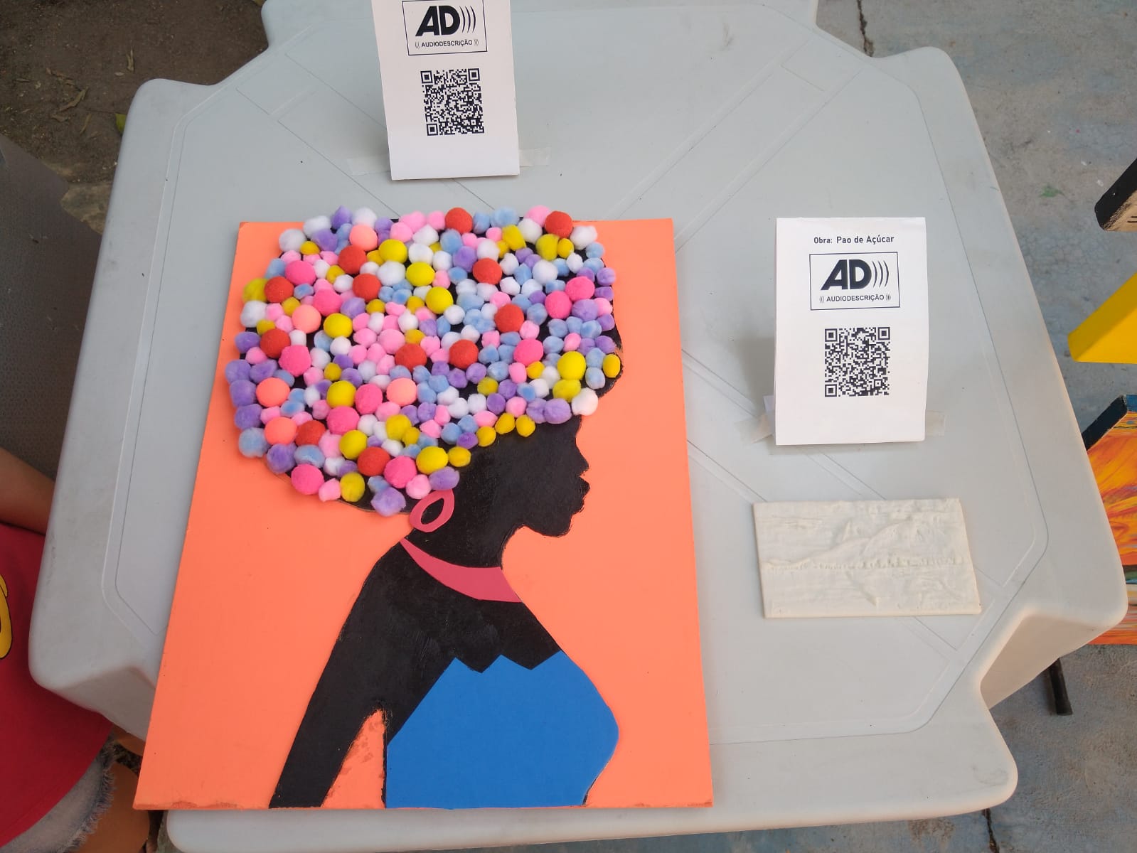 Obras táteis com audiodescrição na Exposição Arte e Conexão 2022: a obra Menina do Black em textura e o relevo do Pão de Açúcar, cada uma com QR Code de audiodescrição